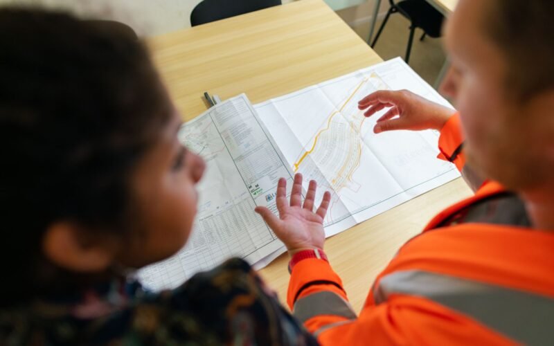 Reflektörlü iş yeleği giyen iki kişi, masadaki teknik çizimi inceliyor